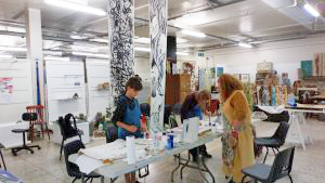 Two Silver Arts Award students, stand at a long table, silk painting guided by artist Wendy Riddick in a large open studio space.