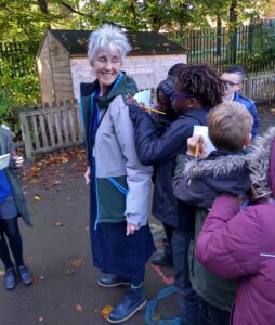 Jean, in a playground with a group of children resting note paper on each other's backs in order to write poetry notes