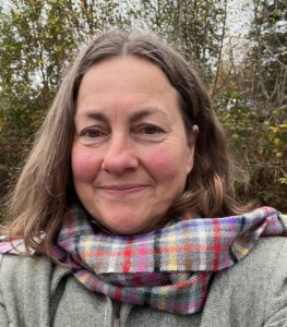 Head & shoulders portrait of a middle-aged white woman with rosy cheeks and mid length, brown hair, wearing a pink & grey tartan scarf and green twill jacket. Trees and grey sky in the background