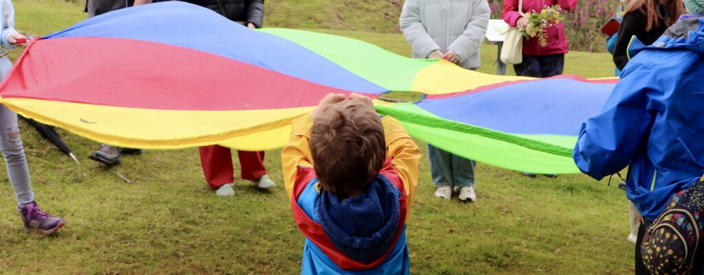 A young child holds a brightly coloured parachute, with other older children and adults