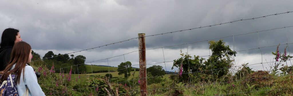 Two people look across the landscape from Oswestry Hill fort, and a cloudy grey sky.