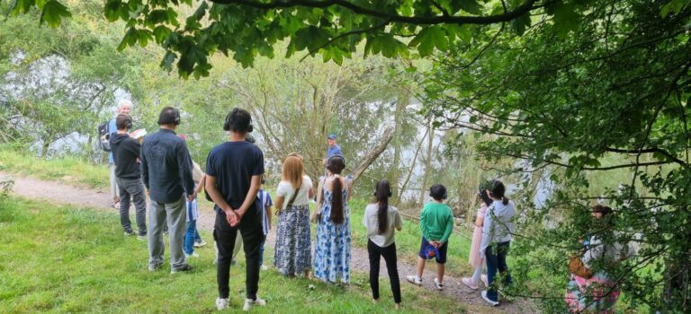 A group of people wearing headphones stand at the rivers edge under leafy trees